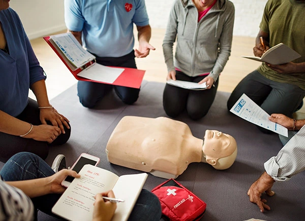 Sesja grupowa podczas kursu kwalifikacyjnego pierwszej pomocy w pracy z uczestnikami robiącymi notatki i przeglądającymi procedury pierwszej pomocy wokół manekina CPR